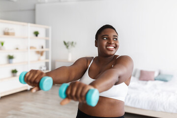 Smiling curvy young black woman doing exercises with dumbbells, during domestic weightloss training