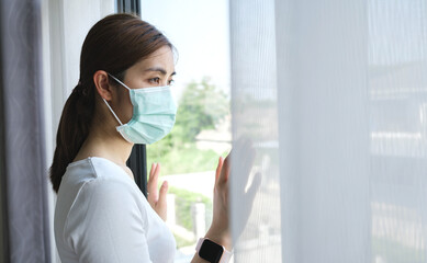 Young woman wearing a medical mask looking through the window. Patient isolated to prevent...