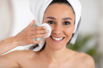 Cheerful young caucasian brunette woman in towel wipes face with cotton pad and lotion in light...