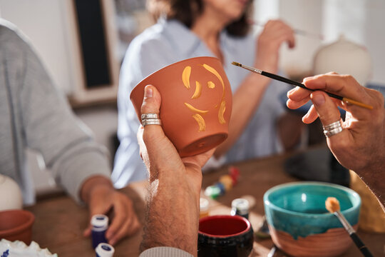 Senior man holding clay pot and brush while drawing patterns on it at the loft studio