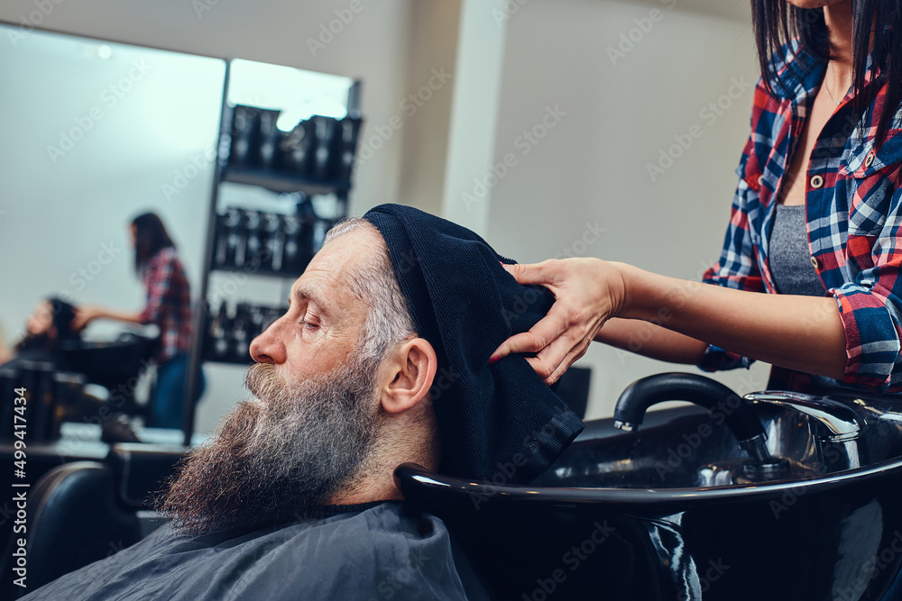 Wall mural shot of bearded elderly man hipster being washing by woman barber in hair salon in daytime.