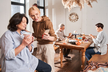 Female teacher holding smartphone and looking with interest at the screen with senior woman