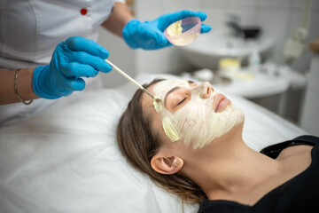 Doctor cosmetologist or dermatologist making face mask in cosmetology salon. Professional Beautician applying face mask on caucasian woman face lying on bed in bathrobe.