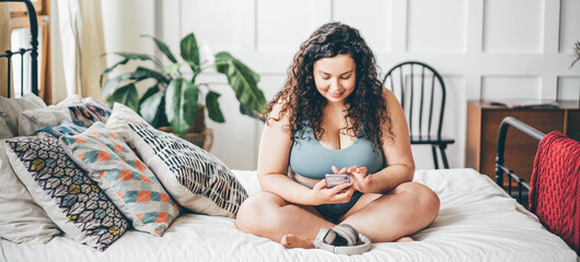 Overweight young woman with loose hair in top and shorts holding smartphone in hand and sitting on double bed.