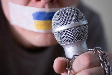 Tape over his mouth with the flag of Ukraine, trying to speak into a microphone.