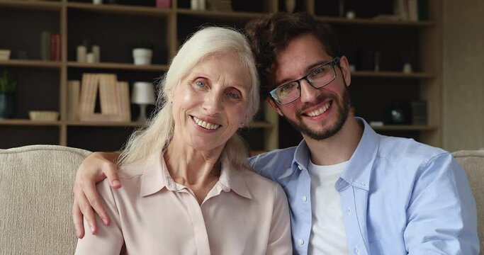 Caring millennial guy in glasses adult child son embrace shoulders of happy older aged female retired mother sit on couch at home look at camera. Bonding senior mom grownup kid smile pose for portrait