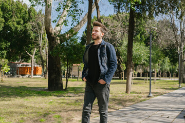 Young man in jeans clothes slowly walking through alley of trees in nice day at park. Spending time alone in nature. Peaceful atmosphere.