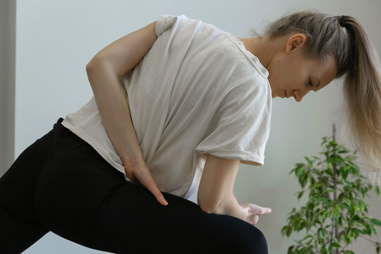 concentrated flexible young woman is doing standing twisting pose, stretching her shoulder and back, wearing beige t-shirt, black leggings and ponytail