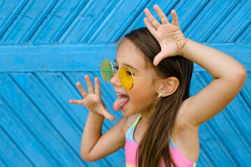 A brunette girl in yellow sunglasses stands against a blue wall of planks and makes a funny grimace, showing her tongue. Naughty ill-mannered child - obrazy, fototapety, plakaty