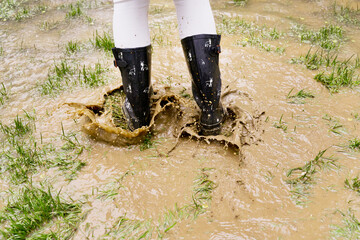 Close-up of unrecognizable woman jumping with galoshes on pond. Horizontal low angle view of wellingtons on rainy pound splashing with mud. Nature and people backgrounds.