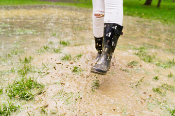 Close-up of unrecognizable woman splashing with galoshes on puddle. Horizontal low angle view of wellingtons on rainy pond splashing with mud. Nature and people backgrounds.
