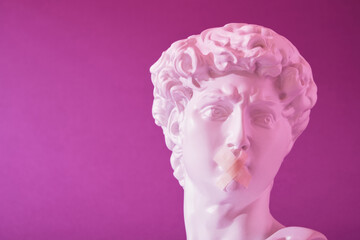 copy of the head of an antique statue of David with a taped mouth in pink neon light on a purple background