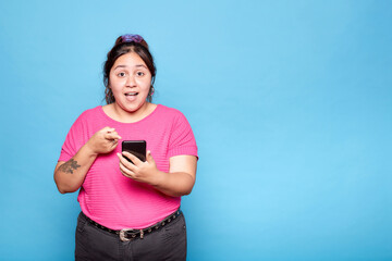 Young curvy Latina woman looking surprised while holding and pointing at her mobile phone, isolated...