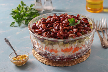 Layered salad Vinaigrette with red beans, pickles, boiled potatoes, beets and carrots in a transparent salad bowl on a blue background