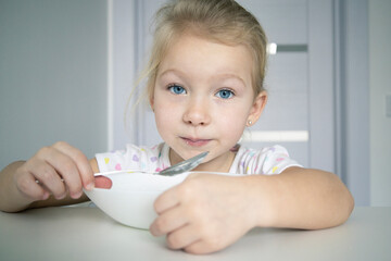 Child girl blonde eats with a spoon from a white plate at a white table.