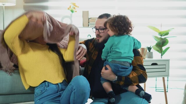 Young couple in their house with a small baby spend time playing. Mom makes the baby laugh by hiding under the blanket