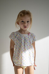 Frightened child girl blonde stands against the background of a white wall.