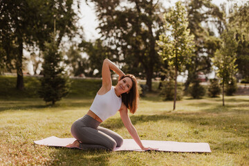 Fototapeta premium full length yoga concept. sporty beautiful woman with dark hair sit on her legs with raised bent hand on yoga mat outdoors at park background in sunset. 