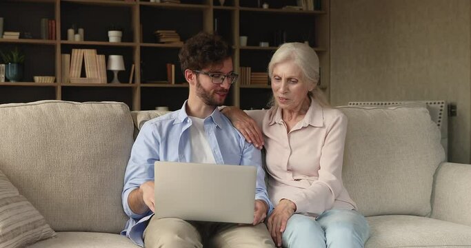 Happy adult son grandson sit on couch register mature mother grandma at social media successfully using laptop. Excited young guy teach older mom to play online game enjoy victory together with parent