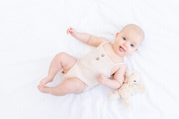 a newborn baby baby girl in a cotton suit on a white bed at home is lying and smiling with a teddy bear