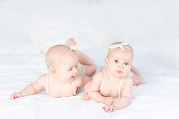 two newborn baby twin girls in a cotton suit on a white bed at home lie on their stomach and smile while playing with each other