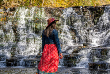 Lady wearing pretty dress, denim jacket and hat standing by a waterfall in the late summer