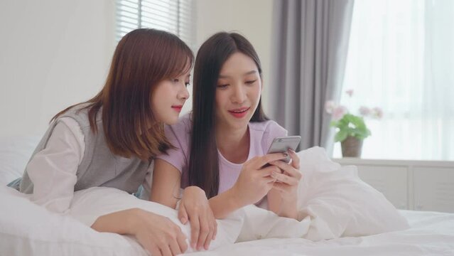 Young beauty Asian women LGBT lesbian happy couple laying on the bed using smartphone together in bedroom. LGBT lesbian couple together indoors concept. Spending nice time at home