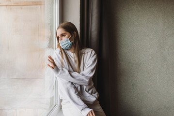 Young woman sitting on the windowsill in the medical mask and looking throw the window. Self isolation and quarantine - obrazy, fototapety, plakaty