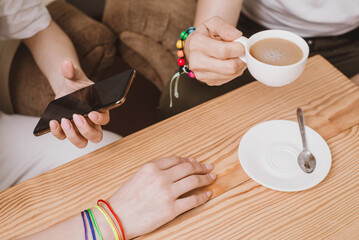 Lesbian couple drinking coffee in a cafe and serfing in smartphone close up. Hands holding rainbow hearts