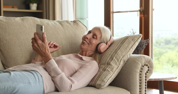 Tranquil aged lady lie on sofa at comfortable pose wear headset choose audio book file music track from phone playlist to spend free time with pleasure. Happy old female use modern tech to enjoy songs