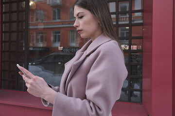 a beautiful girl in a pink coat writes a letter on the go on a smartphone