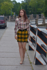 pretty brunette girl in a plaid blouse and skirt on a city street - 499462611