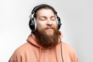 Scandinavian handsome contented man closed his eyes and listens to music in professional headphones isolated on gray background. Happy guy with ginger hairstyle and beard. Modern digital technologies