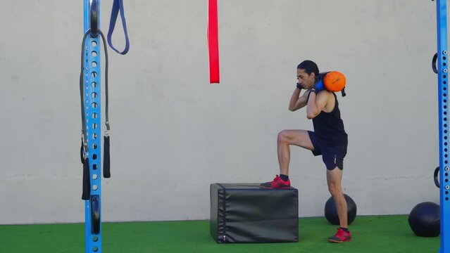 slim young latin man in the outdoor gym doing step up on a box with a sandbag weight. 4k video