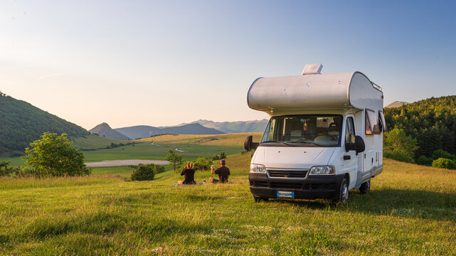 Couple having beer near camper van in Montelago highlands, Marche, Italy. Romantic sunset, unique hills and mountains landscape, alternative vanlife vacation concept.