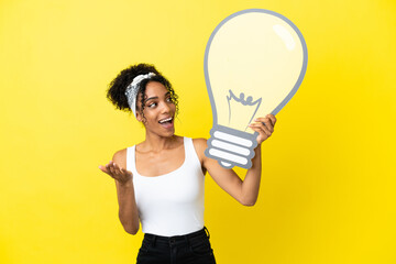 Young african american woman isolated on yellow background holding a bulb icon with surprised...