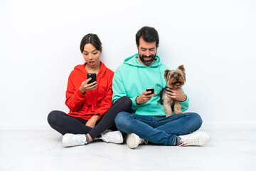 Obraz na płótnie Canvas Young caucasian couple sitting on the floor with their pet isolated on white background Surprised and sending a message or email with the mobile