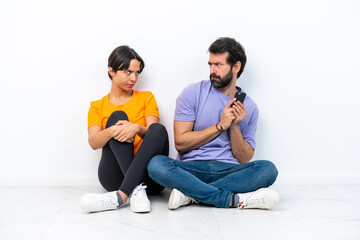 Young caucasian couple sitting on the floor isolated on white background reading a message with the mobile