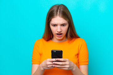 Fototapeta premium Teenager Russian girl isolated on blue background looking at the camera while using the mobile with surprised expression