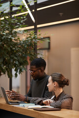 Vertical warm toned portrait of two young colleagues discussing work at standing desk in modern...