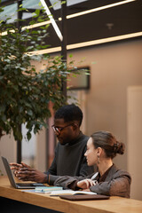 Vertical warm toned portrait of two young colleagues discussing work at standing desk in modern office