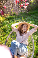 Young beautiful happy smiling woman in a hat enjoying spring while sitting in a blooming garden
