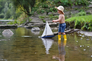 Happy little kid boy in yellow rain boots playing with paper ship boat by huge puddle on spring or...