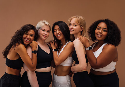 Five women of different body types embracing together. Cheerful females in sport clothes posing on brown background.