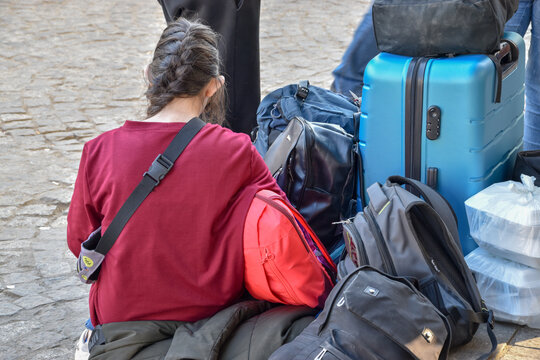 A girl from Ukraine arrived with her family at a train station in Poland. A girl with a braid is sitting on a bag on the street.