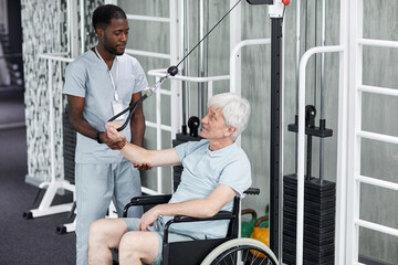 High angle portrait of African American rehabilitation therapist assisting senior man in wheelchair doing exercises in gym at healthcare clinic