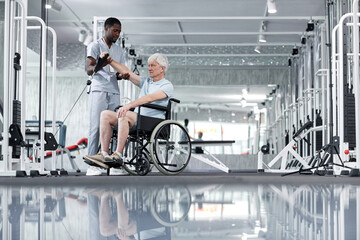 Wide angle view at white haired senior man using wheelchair in gym with rehabilitation therapist assisting, copy space