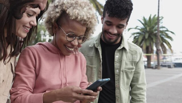 Happy diverse student friends laughing together while watching funny videos on smart phone - Young multiracial people having fun using social media mobile app