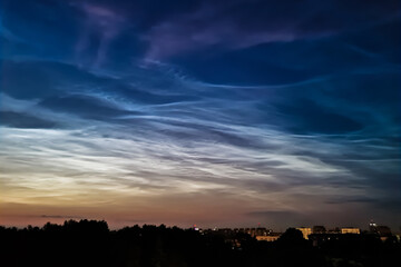 Noctilucent clouds in night sky. Rare atmospheric phenomenon over city. Shining clouds over silhouette of night town.