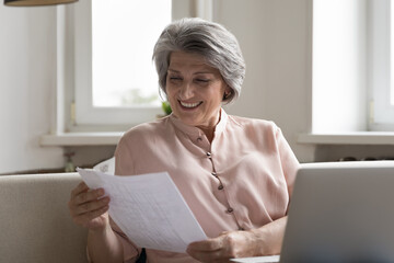 Smiling happy attractive older woman holds paper bills or letter enjoy good news in legal notice...